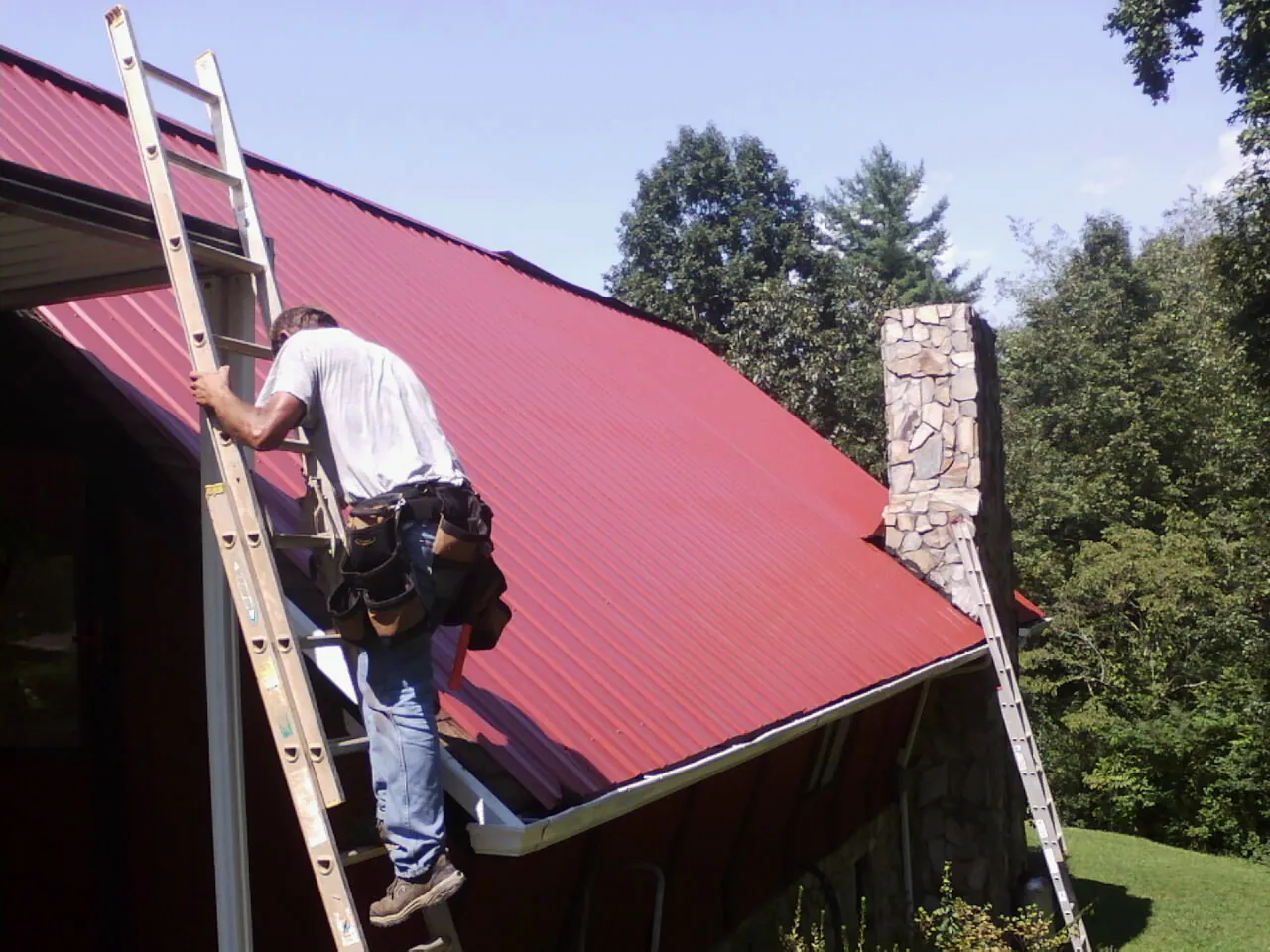 Roofer on a ladder beside a red metal roof and stone chimney — Soffit & Fascia Repair by Roofing Brothers in Stockholm